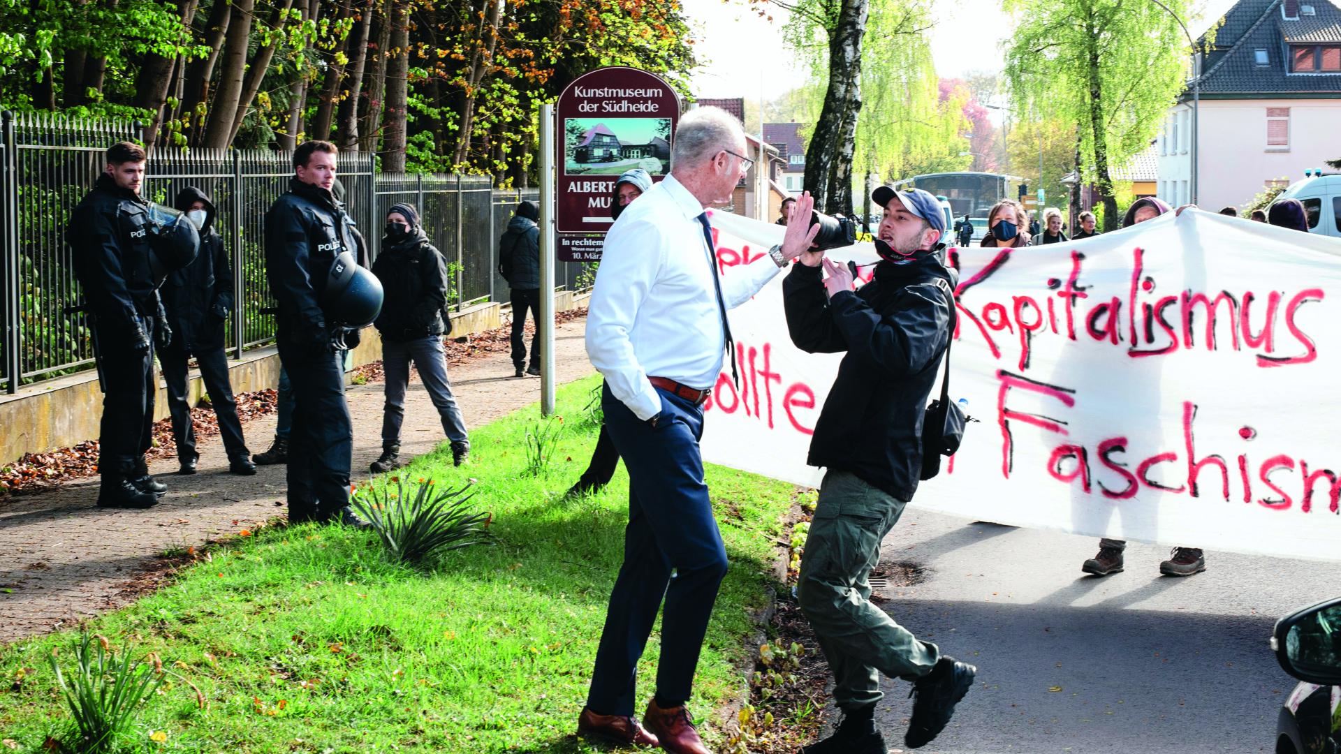 Ein Mann mit Hemd und Krawatte fasst einem Journalisten mit der Hand in seine Kamera. Dahinter stehen Menschen und halten einen weißen Banner mit roter Schrift hoch. Links stehen Polizisten auf einem Weg und schauen in die Richtung des Geschehens.