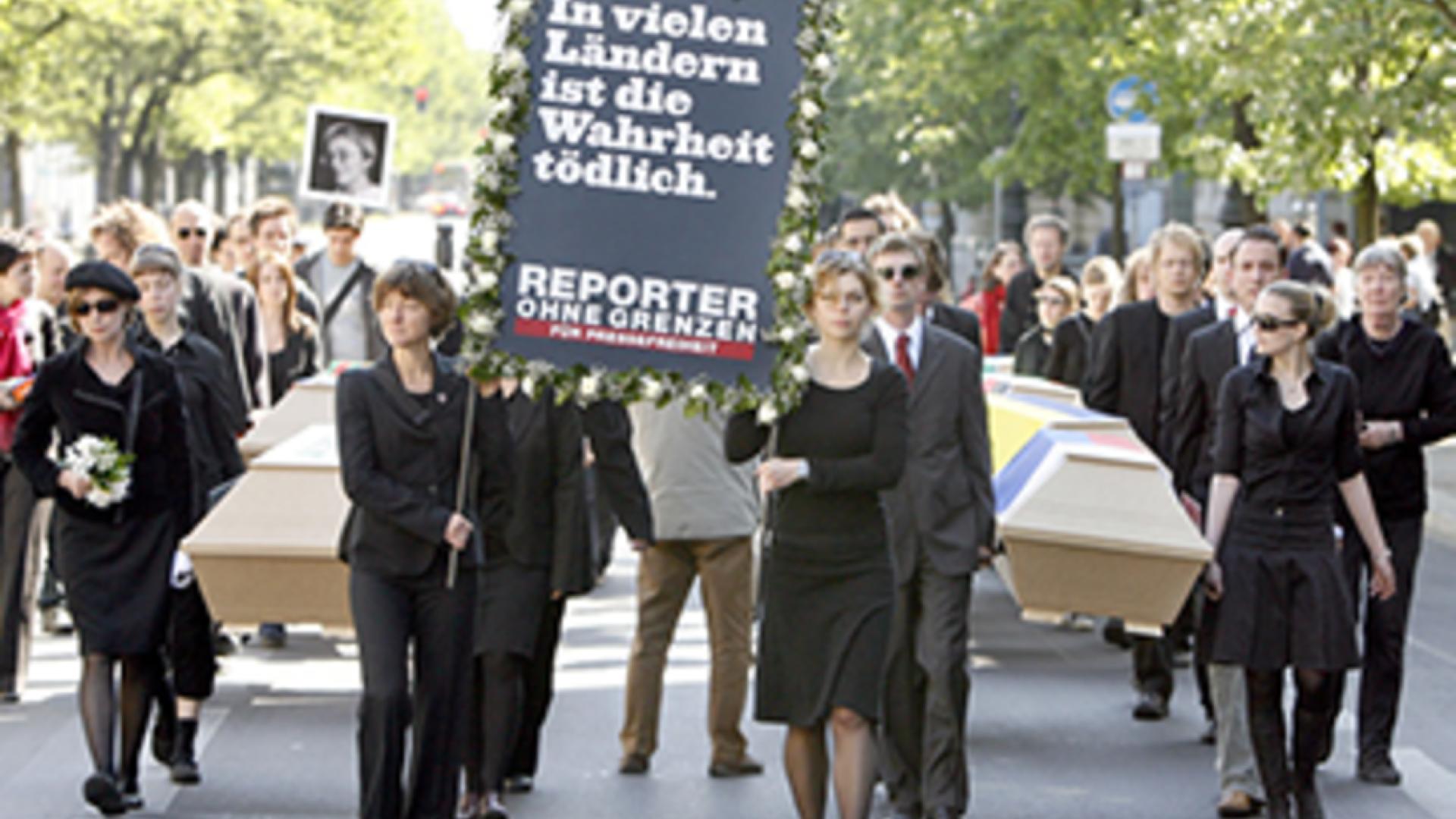 Ein Trauerzug mit mehreren Särgen die von schwarz gekleideten Menschen getragen werden .Eine Person trägt ein Plakat mit der Aufschrift "In vielen Ländern ist die Wahrheit tödlich" in die Höhe hebt