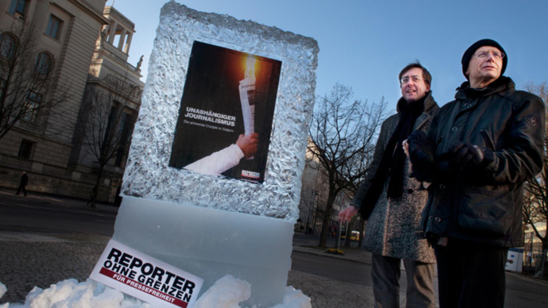 Zwei Männer stehen neben einem Eisblock mit einem Plakat darin: Eine brennende Zeitung mit dem Text „Unabhängiger Journalismus – Die schwerste Disziplin in Sotschi“. Unten das Logo von „Reporter ohne Grenzen – Für Pressefreiheit“.