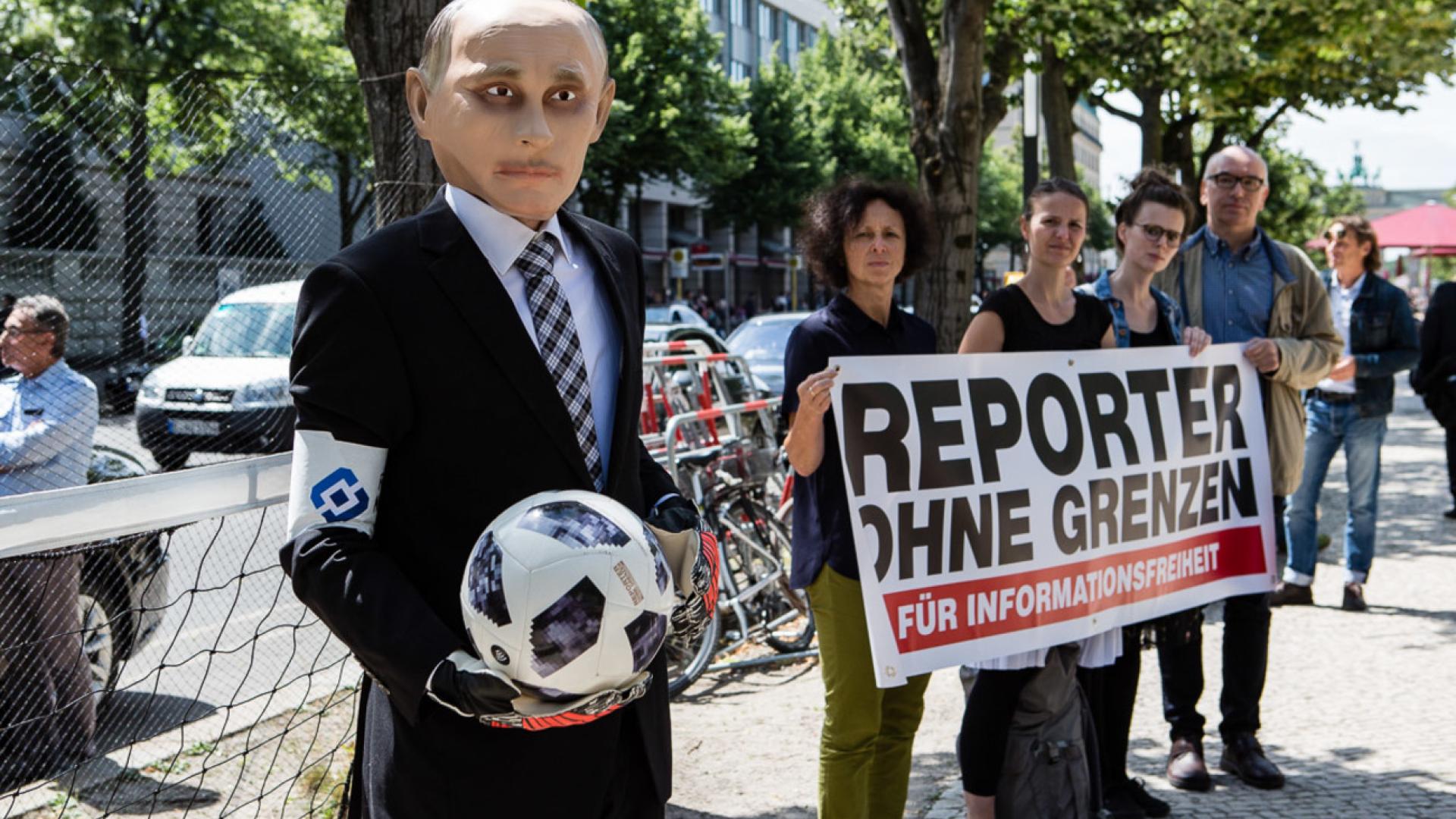 Ein als Vladimir Putin verkleideter Mensch trägt eine Kapitänsbinde und hält einen Fußball in der Hand. Hinter ihm steht ein kleines Tor, neben ihn eine Gruppe Menschen mit dem Logo von ROG.