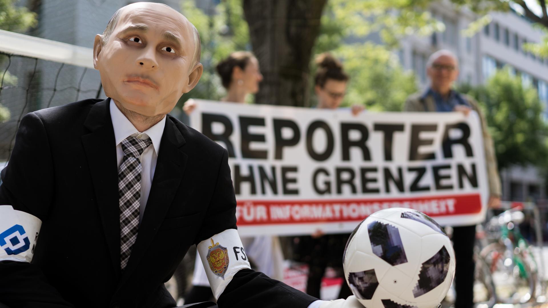 Ein als Vladimir Putin verkleideter Mensch steht vor einer Gruppe die ein Banner von Reporter ohne Grenzen halten. Er trägt einen Torwarthandschuh und hält darauf einen Ball