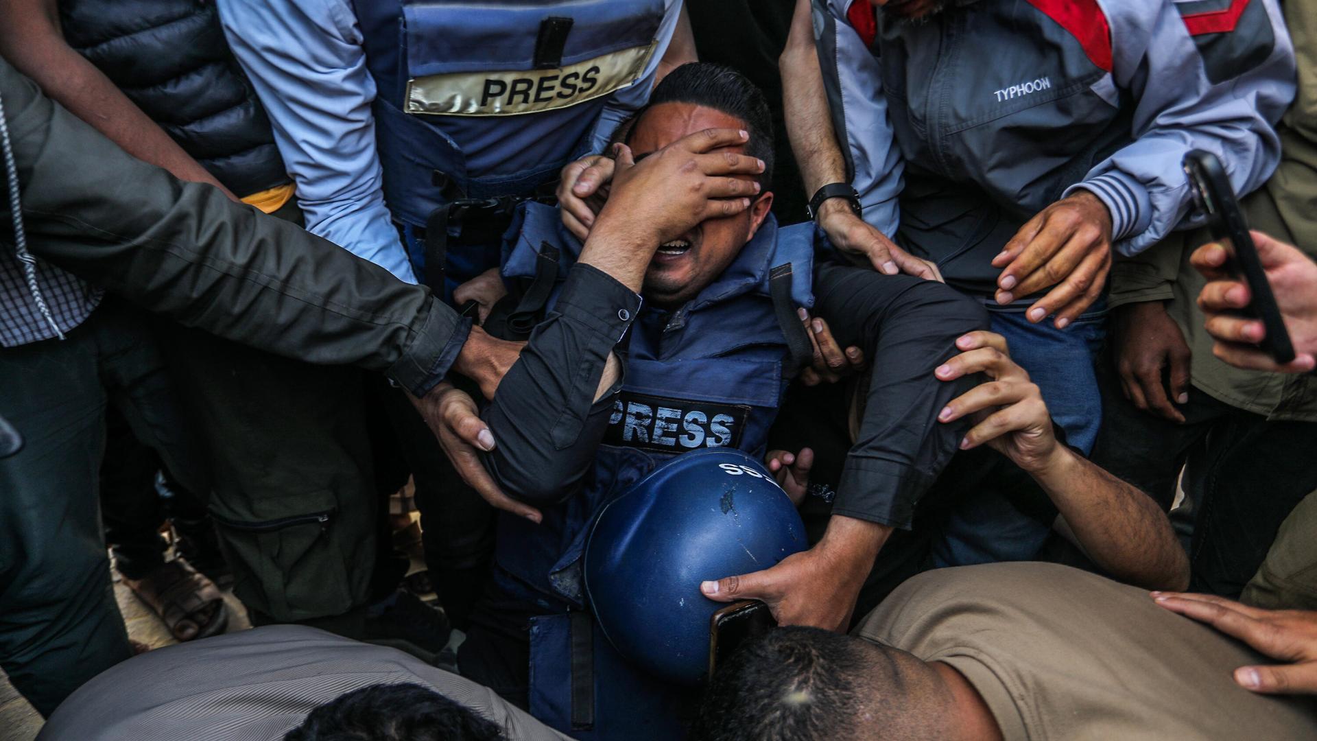 Ein palästinensischer Journalist trauert. Er ist auf die Knie gefallen und hält sich die rechte Hand vor das Gesicht. Er trägt eine Schutzweste und hält einen blauen Helm in der anderen Hand. Um ihn herum sind andere Journalisten, die ihn halten.