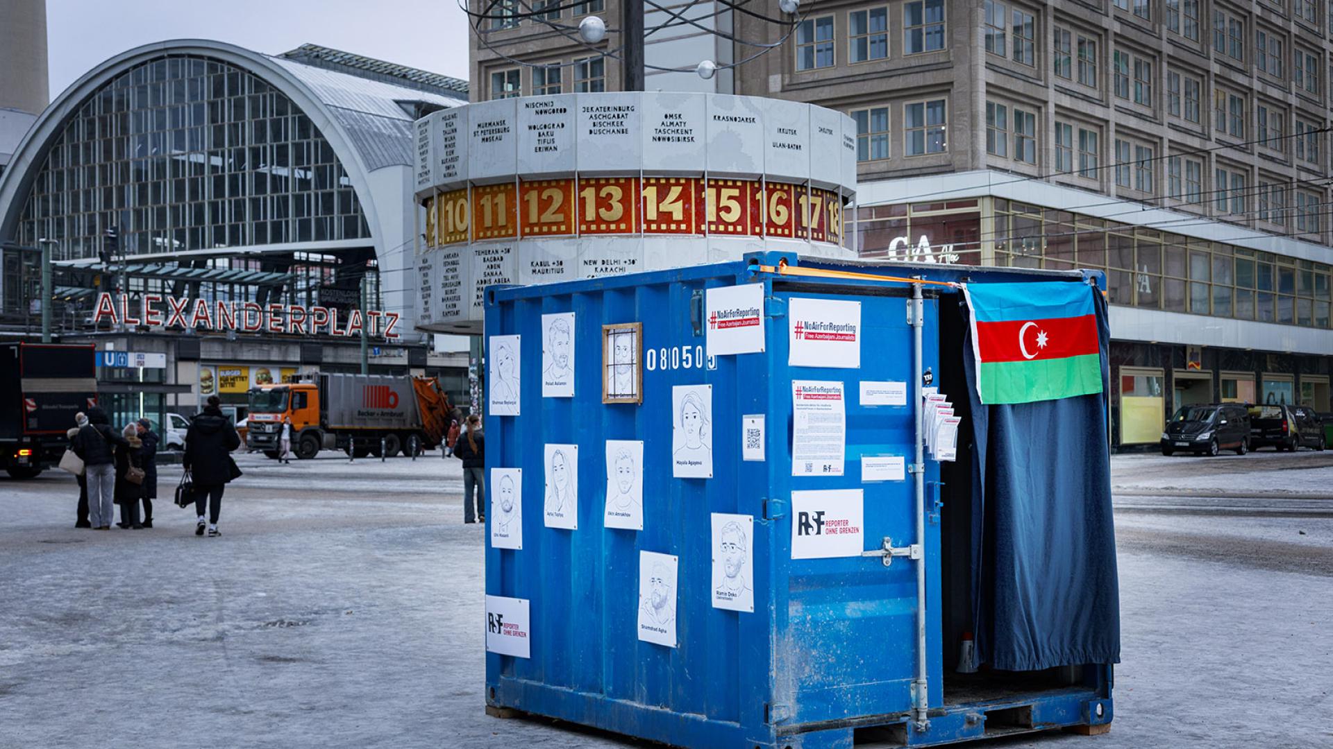 Ein mit Illustrationen und der aserbaidschanischen Flagge behängter blauer Container, der die Haftbedingungen von Journalist*innen in Aserbaidschan abbildet. Im Hintergrund die Weltzeituhr und der Bahnhof Alexanderplatz.