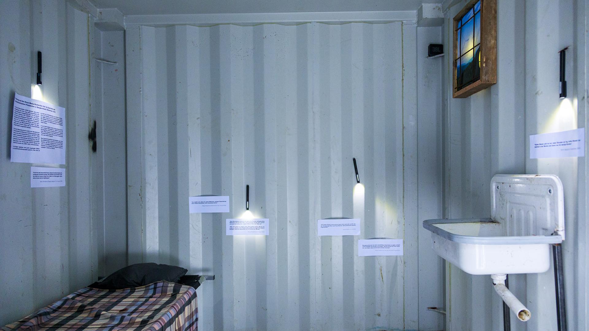 Dunkler Innenbereich eines Containers, rechts ein dreckiges Waschbecken. Links ein Feldbett. Auf den Containerwänden hängen Zitate und Berichte der inhaftierten Journalist*innen über die Haftbedingungen.