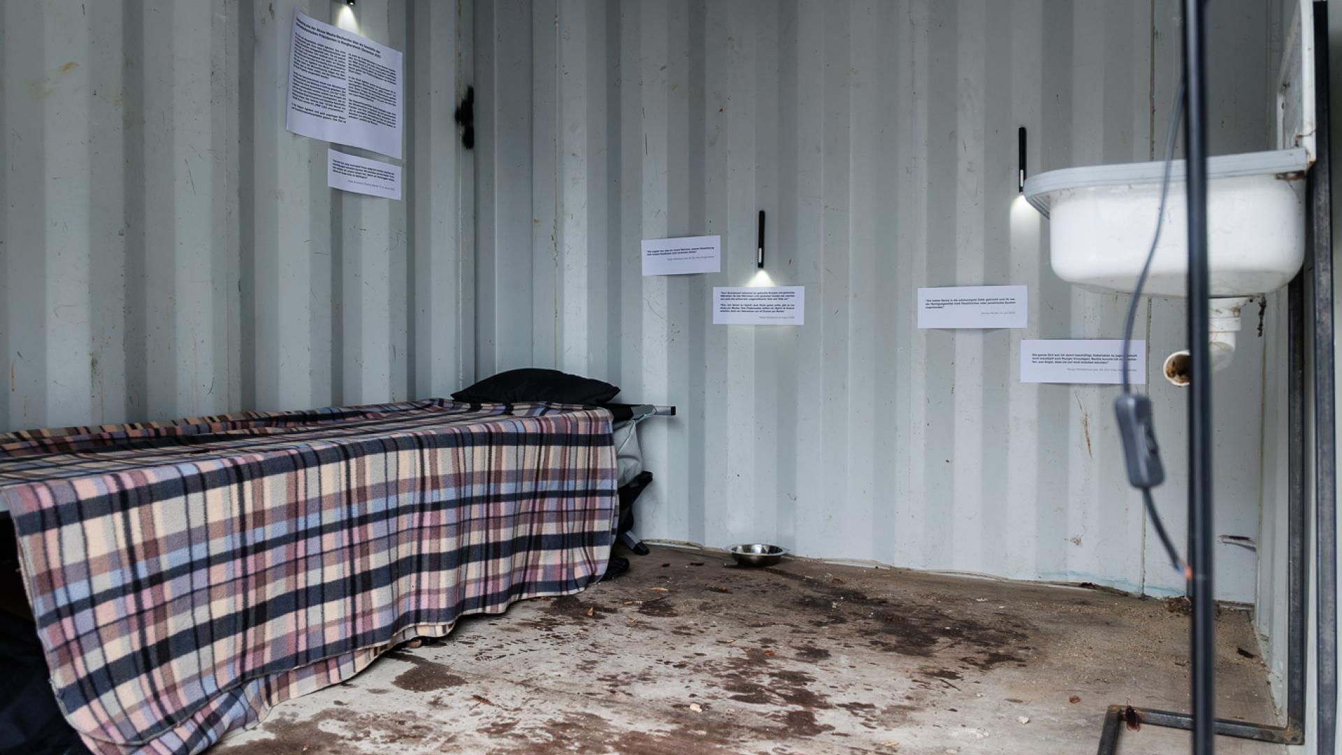 Dunkler Innenbereich eines Containers, rechts ein dreckiges Waschbecken. Links ein Feldbett. Auf den Containerwänden hängen Zitate und Berichte der inhaftierten Journalist*innen über die Haftbedingungen.
