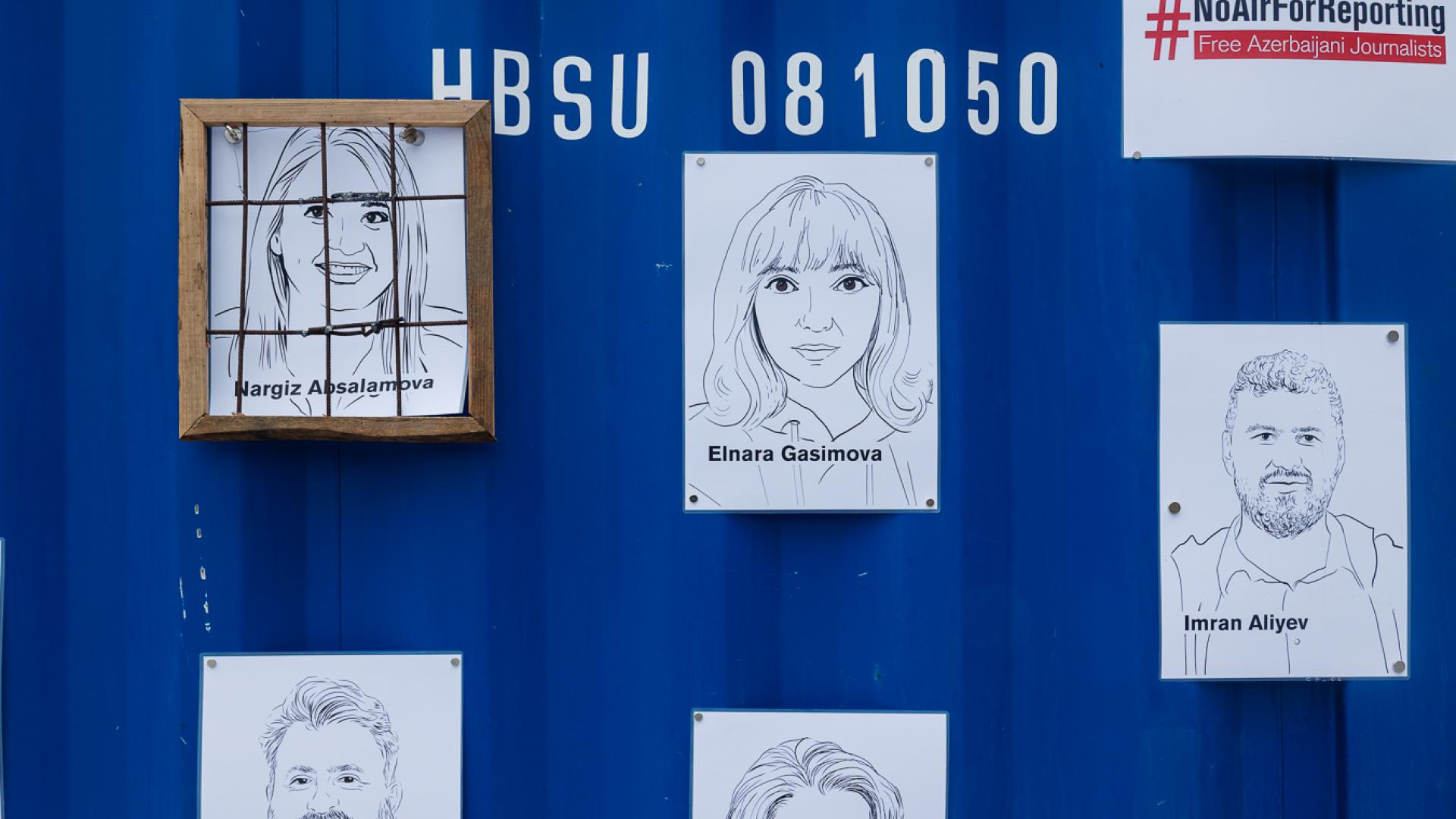 Außensicht auf Containerwand, mehrere Illustrationen von inhaftierten Journalist*innen, eine Illustratien umhängt von nachgebautem Fensterrahmen mit Gittern, symbolisch für ein Gefängnis stehend.