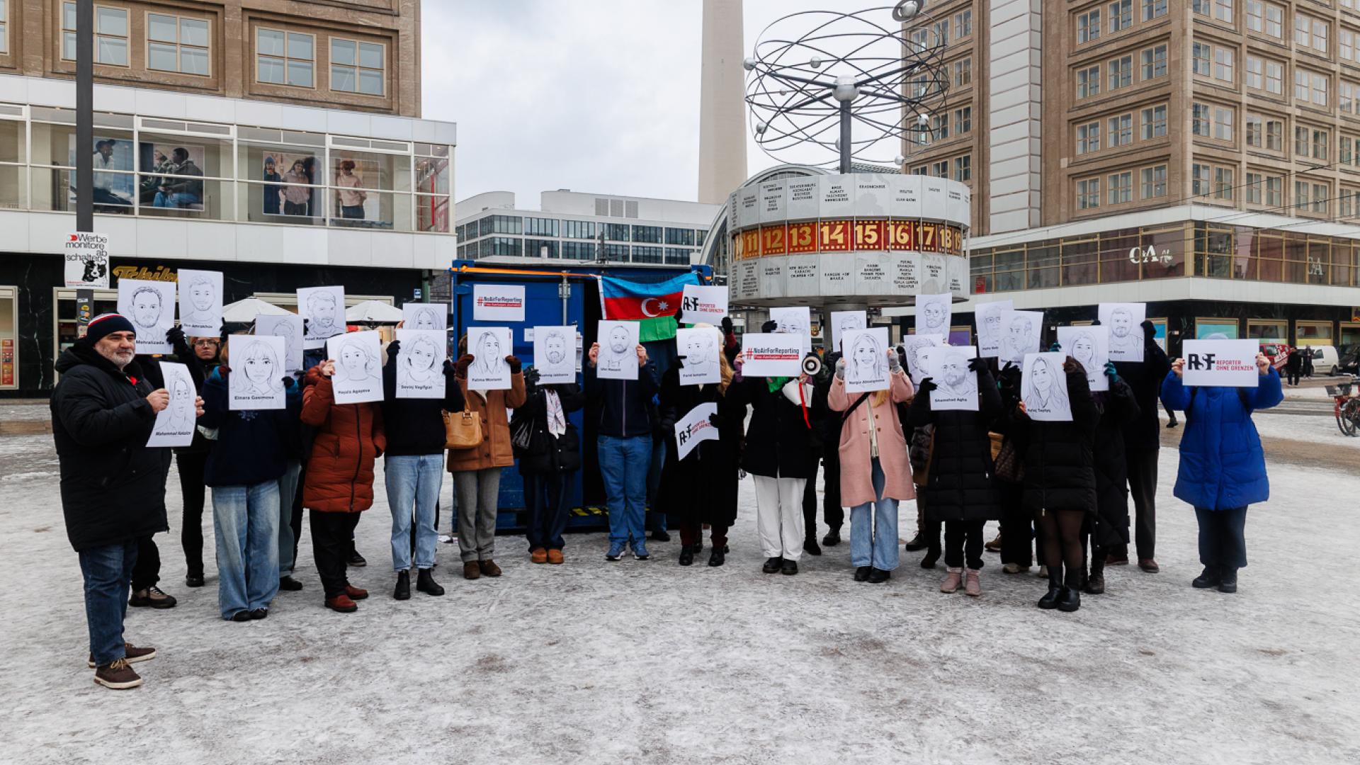 Kundgebung mit etwa 25 Personen auf Alexanderplatz vor blauem Container mit Aserbaidschan-Fahne. Die Menschen halten vor ihren Gesichter Illustrationen von inhaftierten Medienschaffenden in Aserbaidschan.