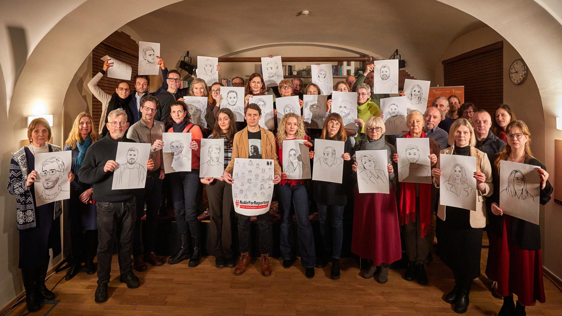 Gruppenfoto mit etwa 25 Personen, die mit Illustrationen von inhaftierten Medienschaffenden in Aserbaidschan ernst in die Kamera gucken.