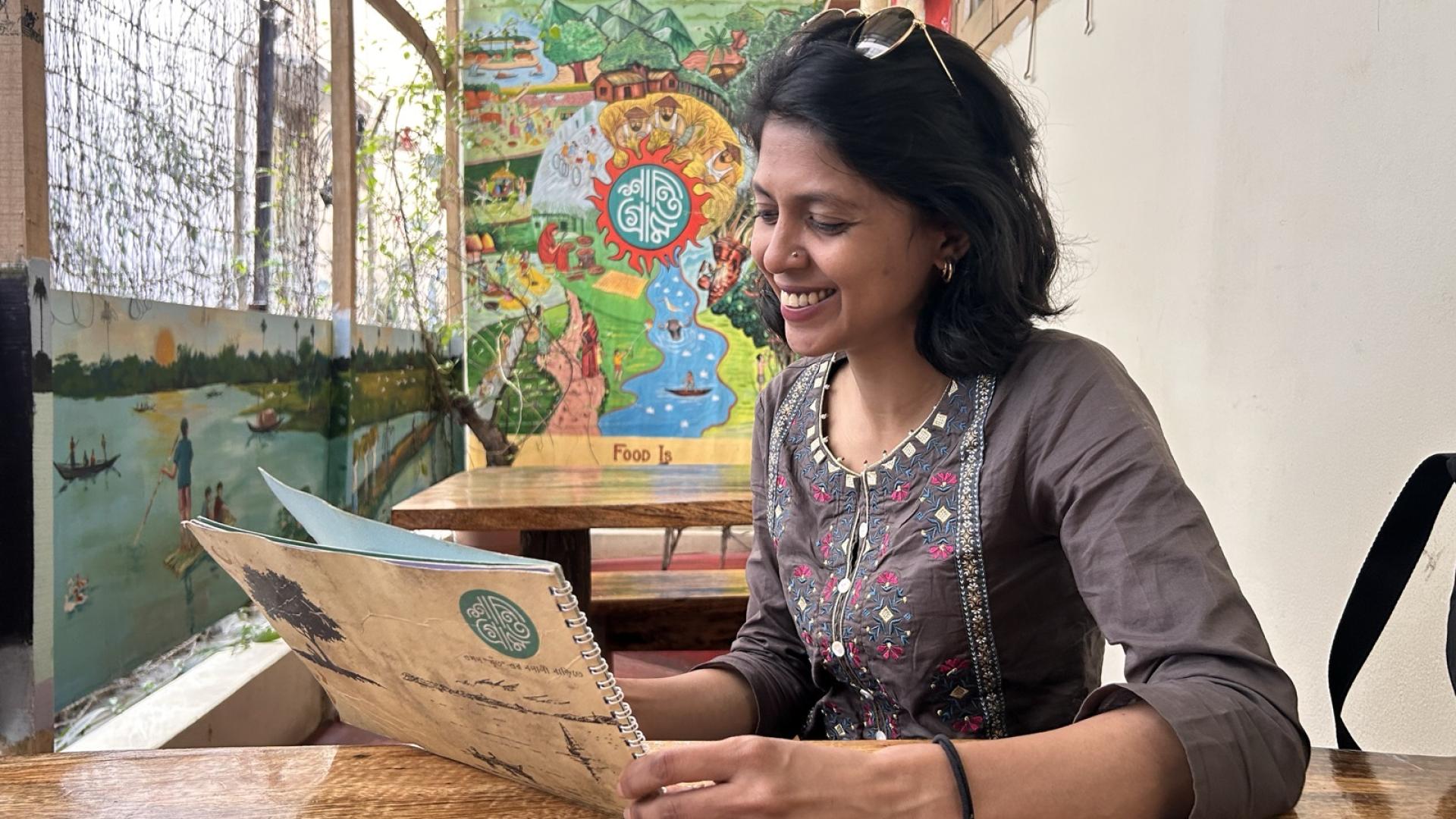 Die Journalistin Shammi Haque sitzt in einem Restaraunt an einem Holztisch und hält die Speisekarte in den Händen. Sie trägt ein braunes Kleid mit aufgestickten Blumen und lächelt.