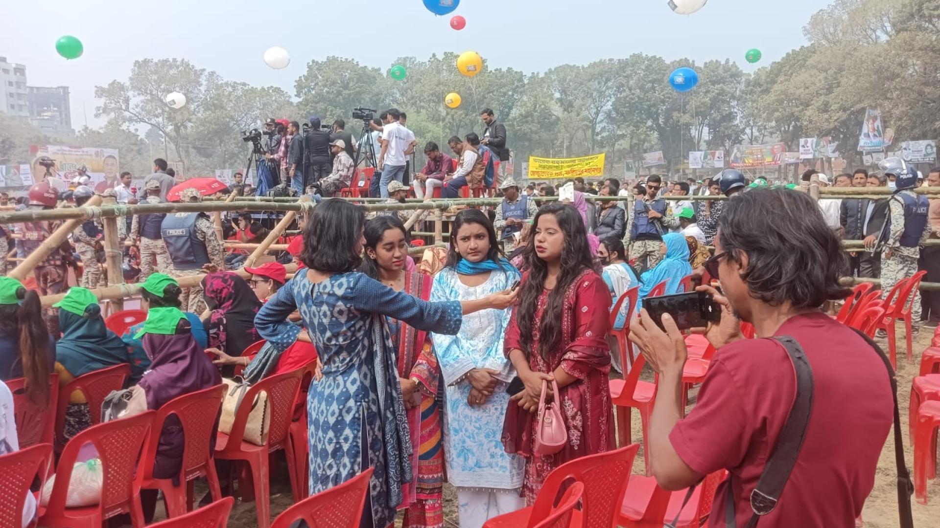 Shammi Haque interviewt eine junge Frau bei einer Wahlkampfveranstaltung der Partei BNP. Um sie herum sind rote Plastikstühle aufgereiht, im Hintergrund sind Menschen und Luftballons zu sehen. 