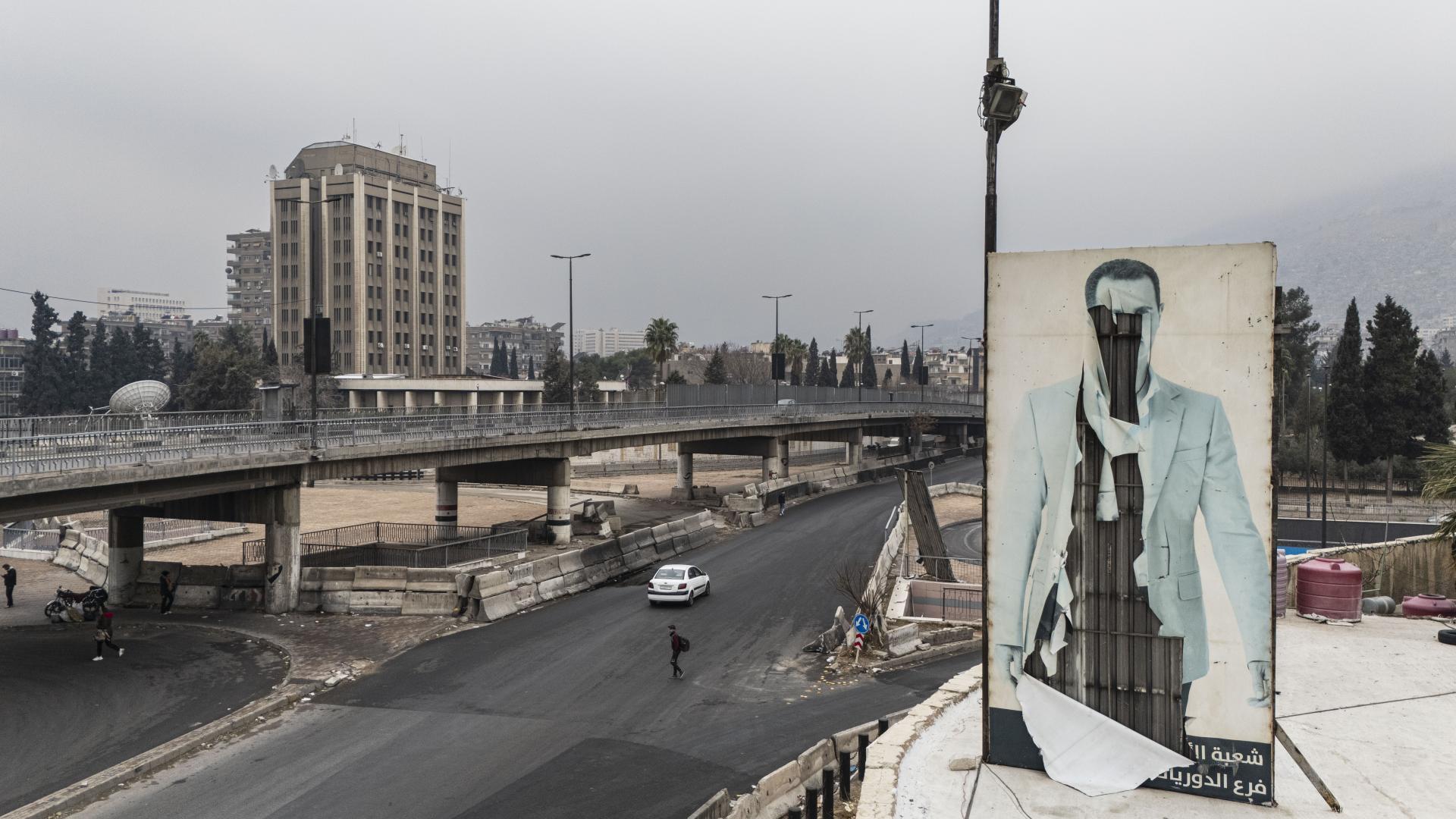 Zu sehen ist eine Straße in Damaskus. Rechts von ihr ist eine zerstörte Plakat Wand des gestürzten Herrschers Assad.