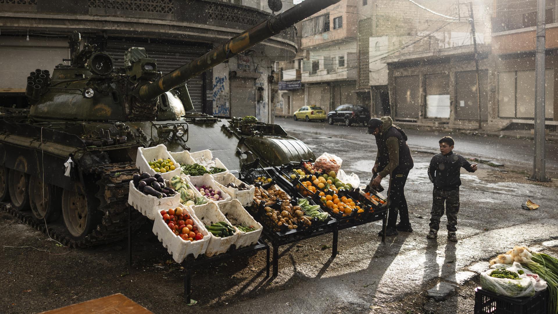 Ein Panzer wurde zu einem Markstand umfunktioniert. Vor ihm lagern nun Obst und Gemüsekisten. Ein kleiner Junge und ein Mann stehen daneben.