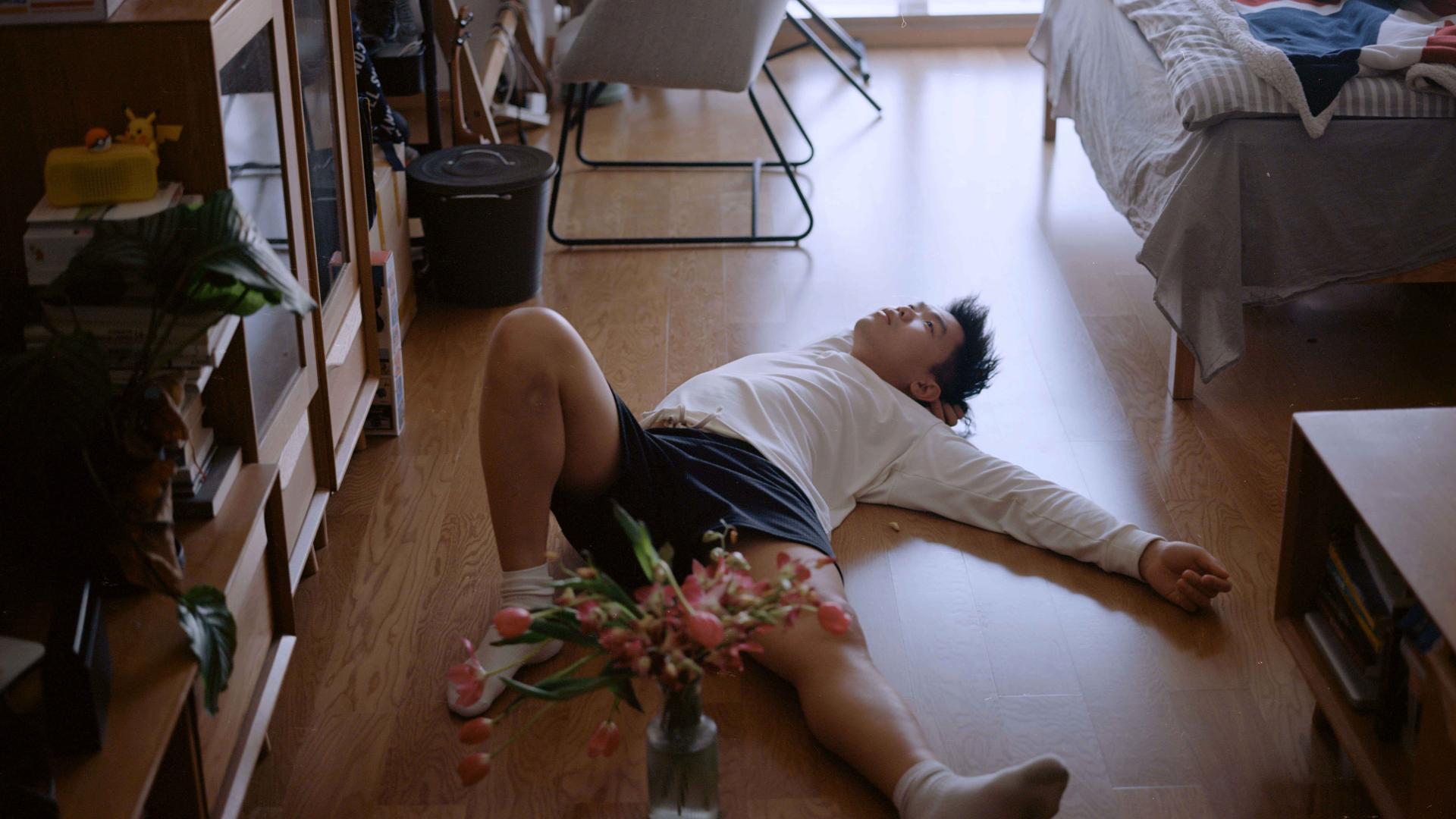 Ein Mann liegt mit von sich gestreckten Armen und Beinen auf dem Boden seiner Wohnung. Vor ihm steht eine Blumenvase mit rosa Blumen.