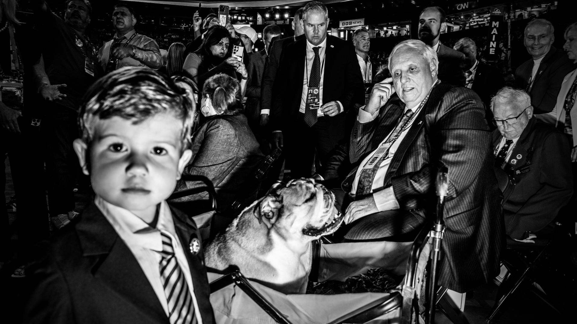 Auf einem schwarz-weiß Foto stehen mehrere Menschen um einen älteren, sitzenden Herren. Vor ihm sitz eine Bulldogge in einem Wagen. Davor steht ein kleiner Junge. Er schaut in die Kamera.