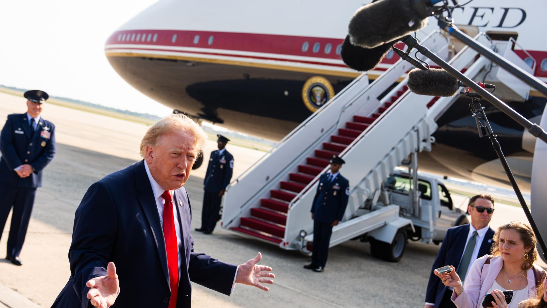 Donald Trump steht vor einem Flugzeug. Dort spricht er mit Journalist*innen, die ihm Mikrofone entgegenhalten. 