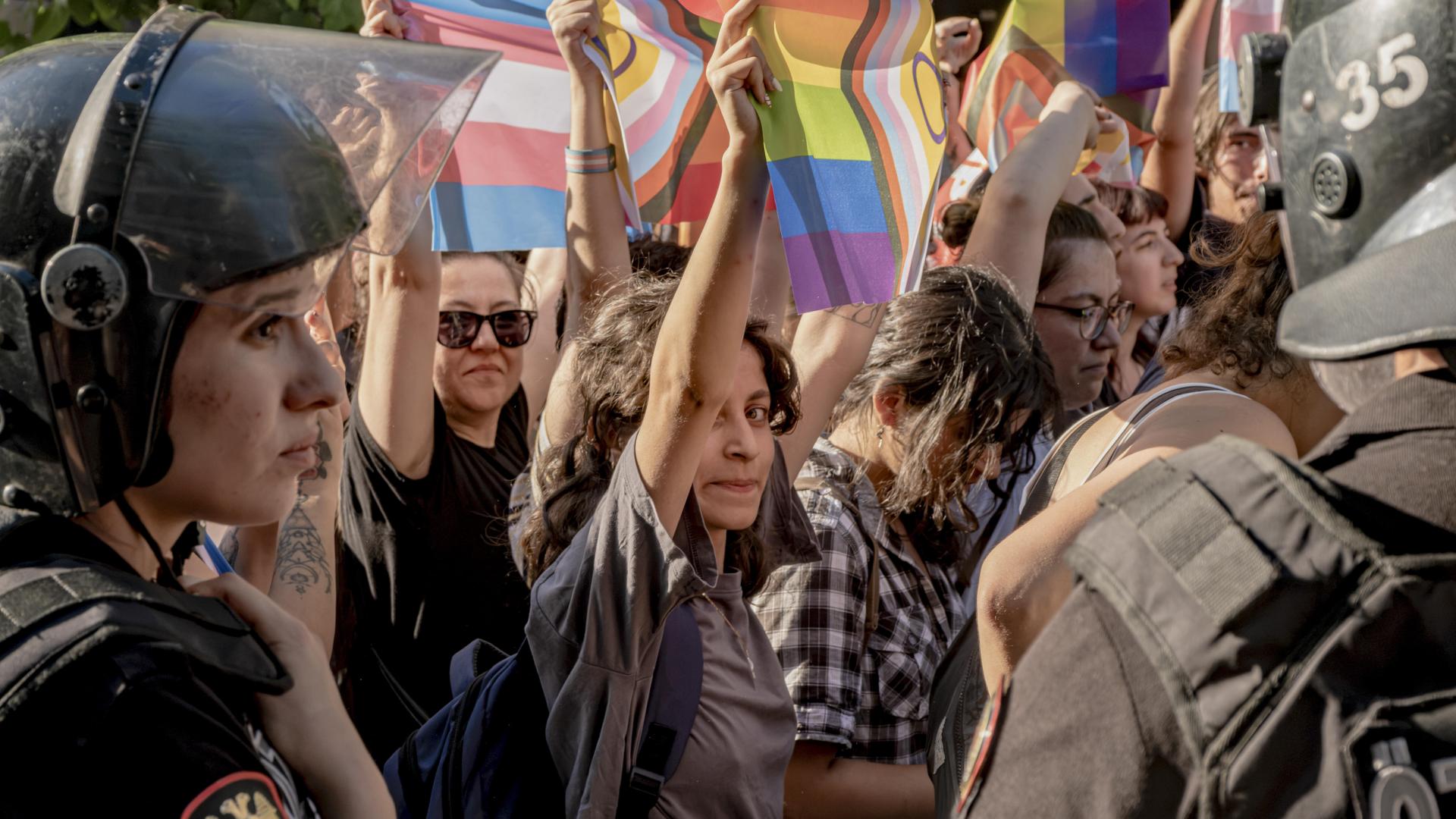 Das Bild zeigt eine Demonstration für LGBTQ Rechte. Eine junge Frau steht im Mittelpunkt und blickt in Richtung Betrachter*in. Ihre Hände sind nach oben gestreckt: Sie hält eine LGBTQ-Flagge. Am Rand befinden sich Polizist*innen in Uniform.