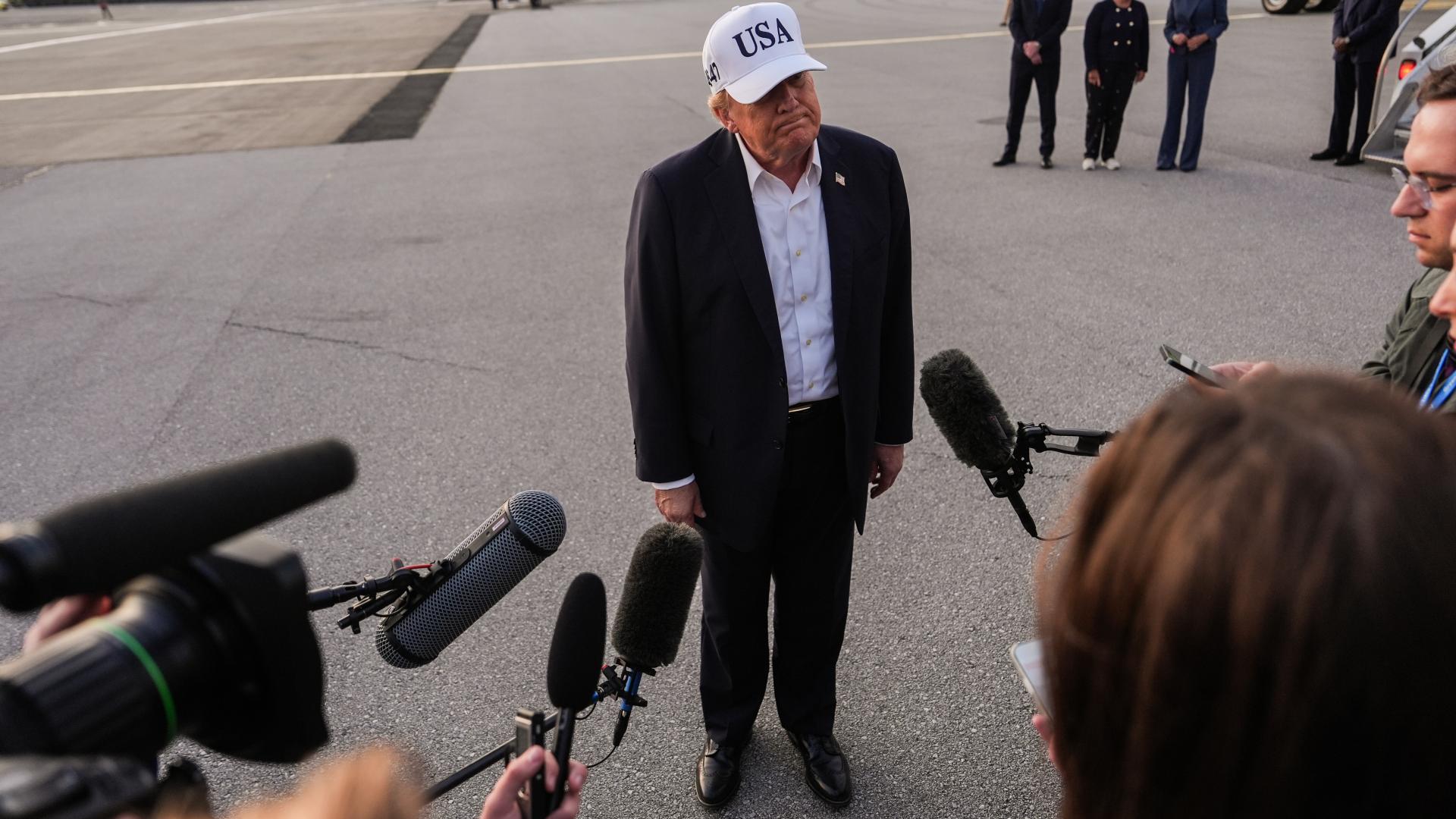 Trump steht im Mittelpunkt des Bildes. Er trägt eine Kappe mit dem Schriftzug "USA". Er ist umrandet von Journalist*innen mit Mikrofonen und Kameras.