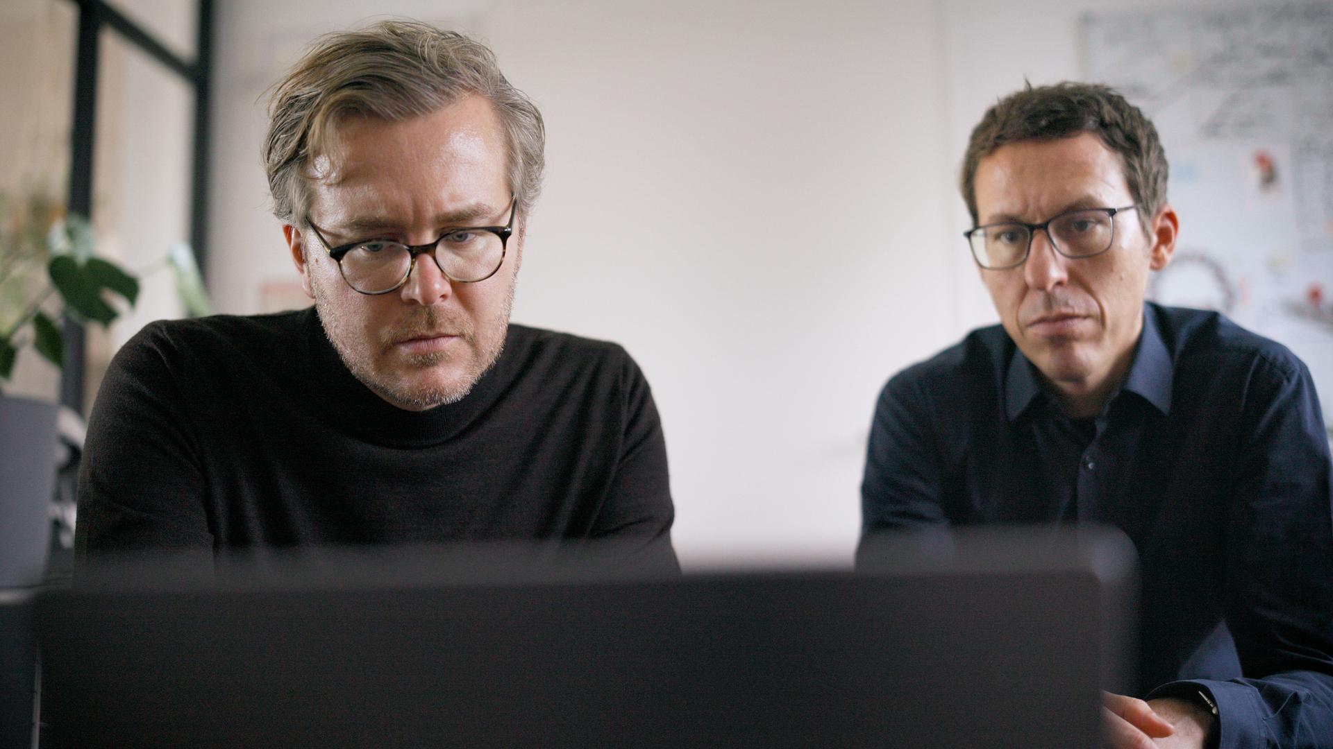 Zwei Männer in dunklen Oberteilen und mit schwarzen Brillen sitzen vor einem Laptop, im Hintergrund eine weiße Wand und eine Pflanze.