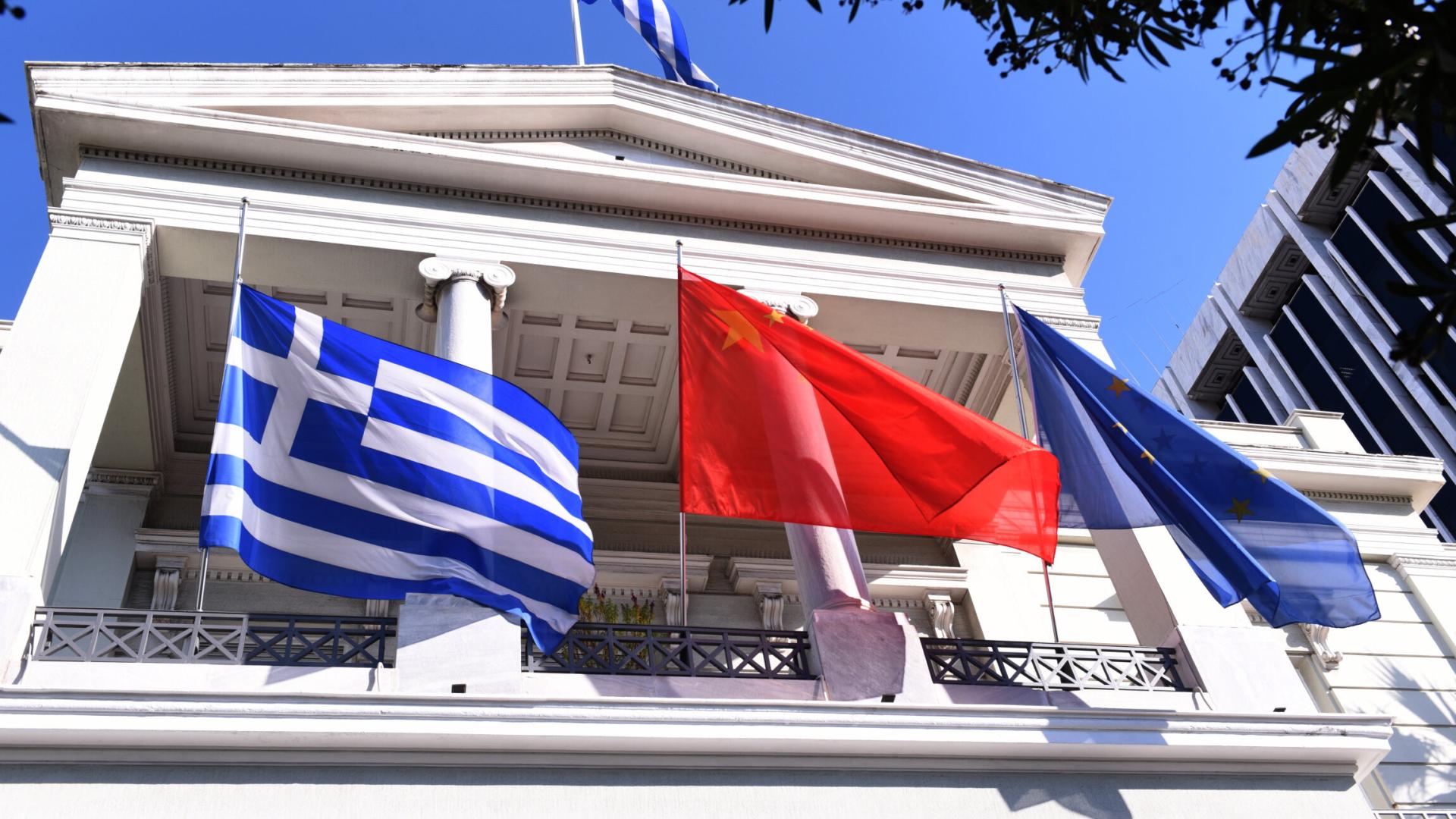 Drei Flaggen vor dem griechischen Außenministerium: links die griechische, in der Mitte die chinesische und rechts die Flagge der EU. Im Hintergrund steht die Botschaft, ein weißes Haus im altgriechischen Stil mit klassischer Architektur.